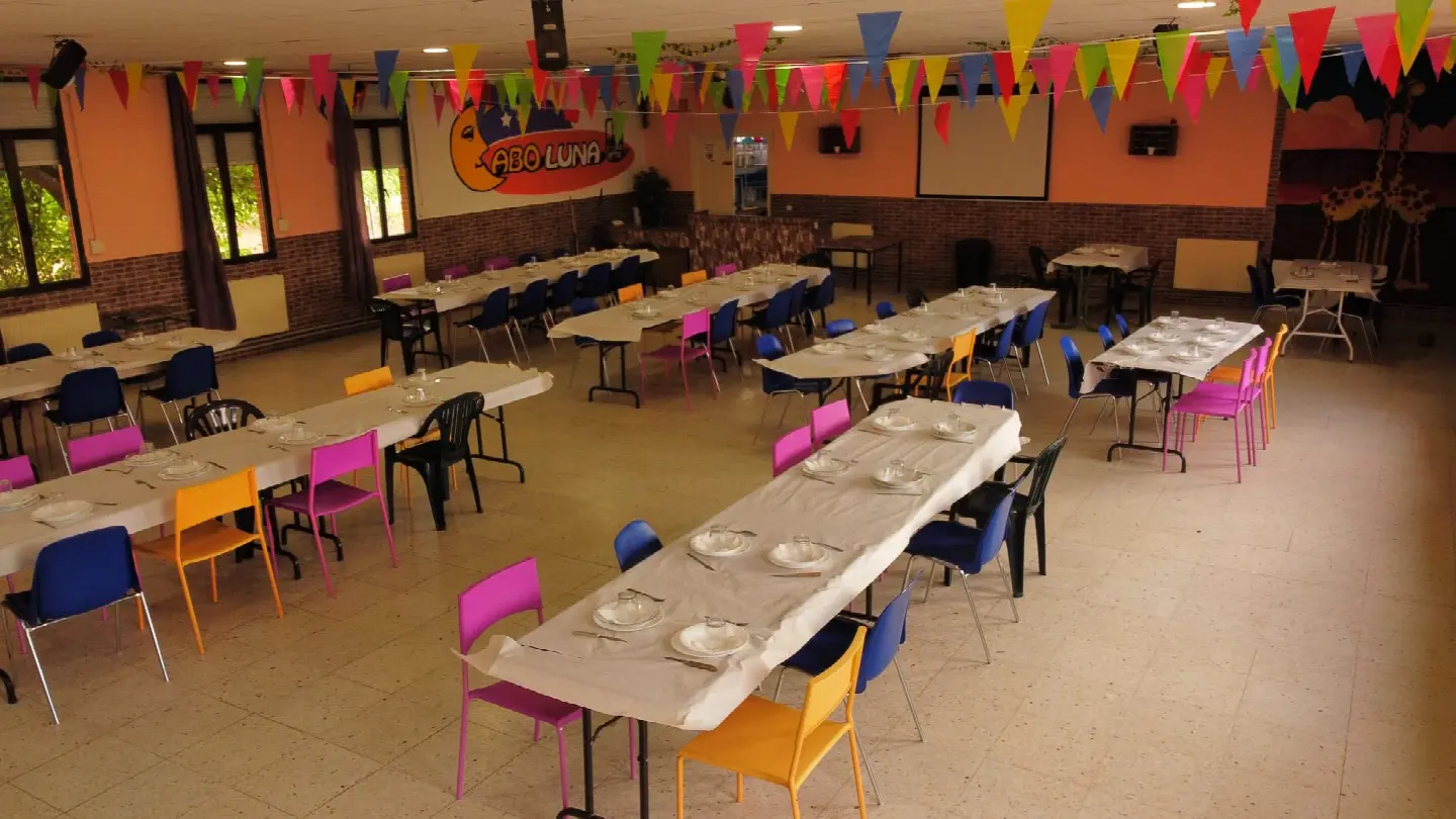 Comedor montado para un grupo: mesas largas con mantel, sillas de colores y banderines